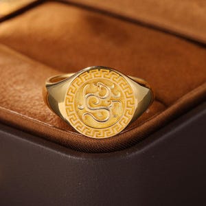 May include: A gold signet ring featuring a circular face with a textured dragon motif in the centre, surrounded by a geometric Greek key border pattern. The ring has a polished gold band and a matte finish on the engraved circular top surface.