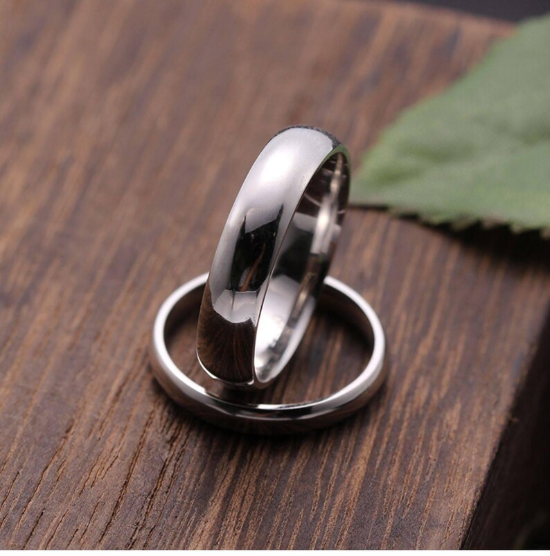 May include: Two polished silver rings, one resting atop the other, against a dark wood grain background. The rings have a smooth, reflective surface, and the top ring is slightly out of focus. The image suggests a theme of commitment or jewelry.