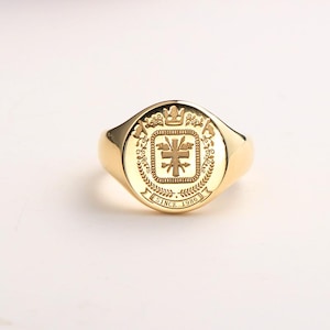 Könnte beinhalten: Ein goldener Siegelring mit einem detaillierten Wappen. Der Ring hat ein rundes Emblem mit einer Krone, floralen Elementen und dem Text "SINCE 1986". Der Ring hat eine polierte, reflektierende Oberfläche.