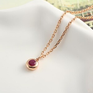 Pode incluir: Um colar em ouro rosa com um pequeno pingente redondo com uma pedra preciosa vermelha escura. A delicada corrente sustenta o pingente, definido em uma moldura simples. O colar é exibido em uma superfície branca.