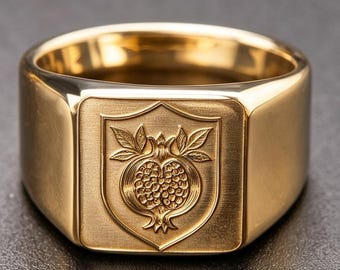 Op de afbeelding: Een gouden zegelring met een plat, rechthoekig vlak met een geborstelde afwerking. Het midden van het vlak toont een gegraveerde schildvorm met een gedetailleerd ontwerp van een granaatappel met bladeren in een gepolijste goudtint.