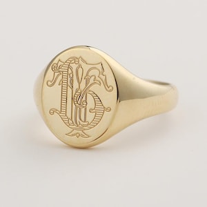 May include: A gold signet ring with an oval face. The face features an engraved monogram design. The ring is made of polished gold and has a smooth, rounded band. The monogram appears to be the letters "J" and "B" intertwined.