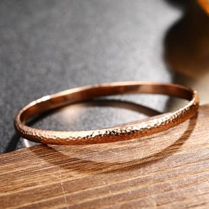 May include: A rose gold-toned bangle bracelet featuring a distinct hammered texture across its entire outer surface. The metal band has a rounded profile and a reflective finish that catches the light, creating highlights along the curved edges.