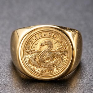 Puede incluir: Un anillo de sello en tono dorado con una cara circular que presenta un diseño en relieve de una serpiente enroscada. La palabra AKARO está inscrita en un semicírculo sobre la serpiente, situada sobre un fondo de paisaje con una montaña.