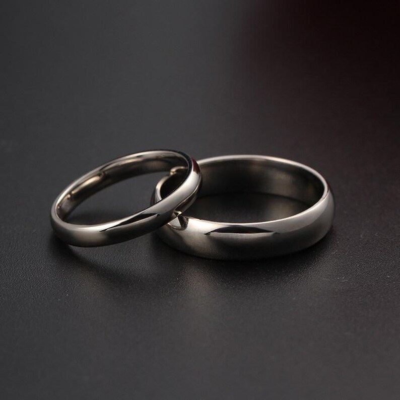 May include: Two silver wedding bands. One band is thin and delicate, while the other is wider with a rounded profile. The rings are positioned together, reflecting light and set against a dark background. The bands are likely made of stainless steel.