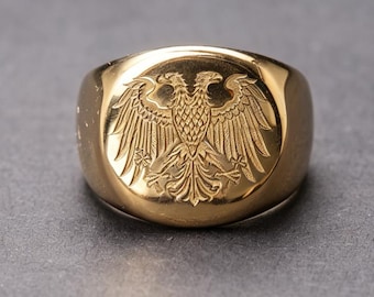 May include: A gold-toned signet ring featuring a deeply engraved double-headed eagle emblem on its flat, circular face. The polished metal surface reflects light, highlighting the intricate feather details and the symmetrical design of the bird.