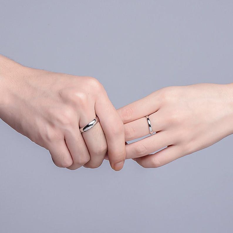 May include: Two silver rings, one with a curved design and the other a simple band, are worn on fingers. The rings are displayed against a soft gray background, highlighting their polished, reflective surfaces. The image suggests a theme of connection or commitment.