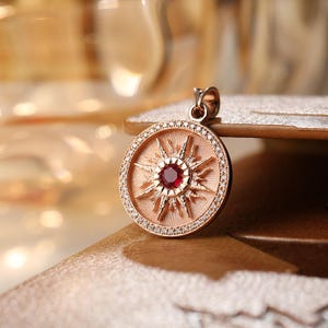 May include: A circular rose gold pendant featuring a central round red gemstone surrounded by a sunburst design. The outer edge of the pendant is lined with small white pavé-set stones, and a small bail is attached to the top for a necklace chain.