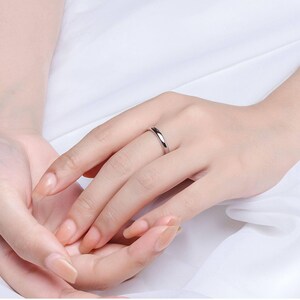 May include: A silver ring is worn on a person's finger. The ring is a simple band with a polished finish. The background is a soft, white fabric. The ring is a classic piece of jewelry.