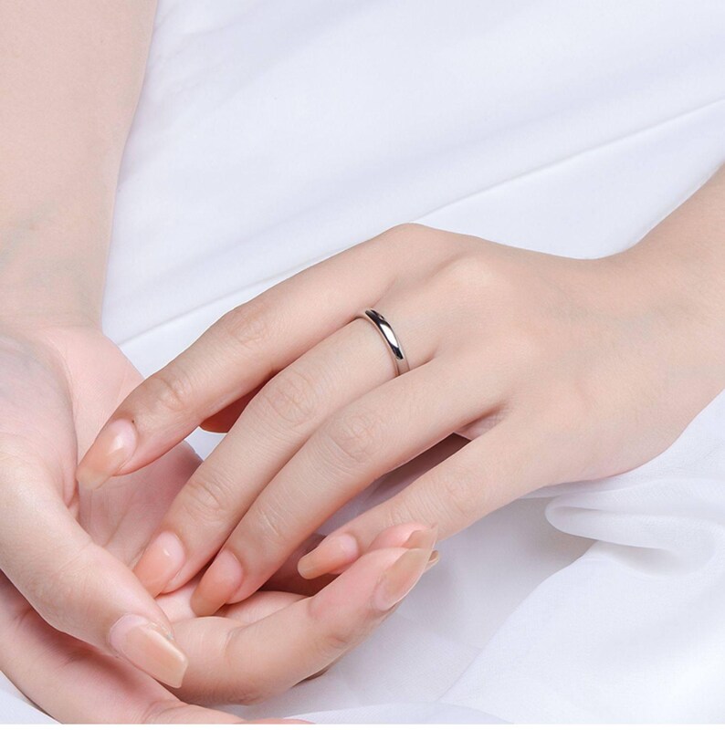 May include: A silver ring is worn on a person's finger. The ring is a simple band with a polished finish. The background is a soft, white fabric. The ring is a classic piece of jewelry.
