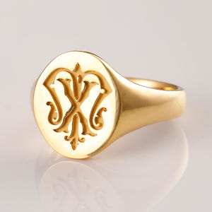 May include: A gold signet ring with an oval face. The face features an engraved design with ornate, stylized letterforms. The ring has a polished finish and a classic, elegant design. The ring is a piece of jewelry.