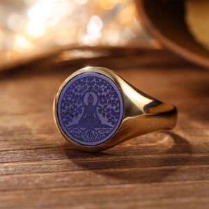 Op de afbeelding: Een goudkleurige ring met een blauwe cirkelvormige inleg met een boom en een zittende figuur. De ring ligt op een houten oppervlak, met een vage achtergrond.