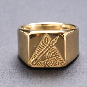 May include: A gold-toned signet ring featuring a flat, square face with an engraved design of a spear positioned diagonally across a textured, triangular pattern. The band is smooth and polished, reflecting light across its metallic surface.