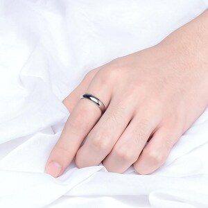 May include: A polished silver ring with a smooth, rounded design. The ring is worn on a finger, set against a backdrop of white fabric. The ring's simple, elegant style is highlighted by the light.