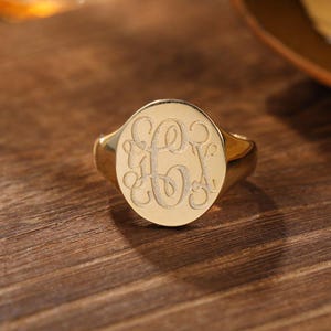 May include: A gold signet ring featuring a circular face with an engraved monogram. The polished metal surface displays three intertwined script letters in a matte finish, creating a distinct contrast against the smooth, reflective gold band.