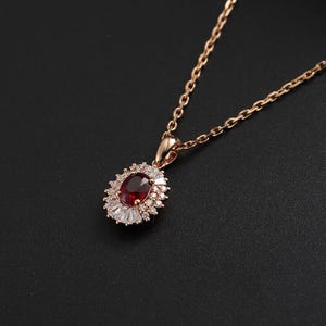 May include: A gold-colored necklace with a pendant featuring a large oval, deep red gemstone surrounded by smaller white gemstones. The pendant is suspended from a delicate chain, set against a dark background.