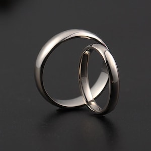 May include: Two silver wedding bands, one larger and one smaller, interlocked against a dark background. The rings have a polished, reflective surface. The smaller ring has the marking "950" on the inside.