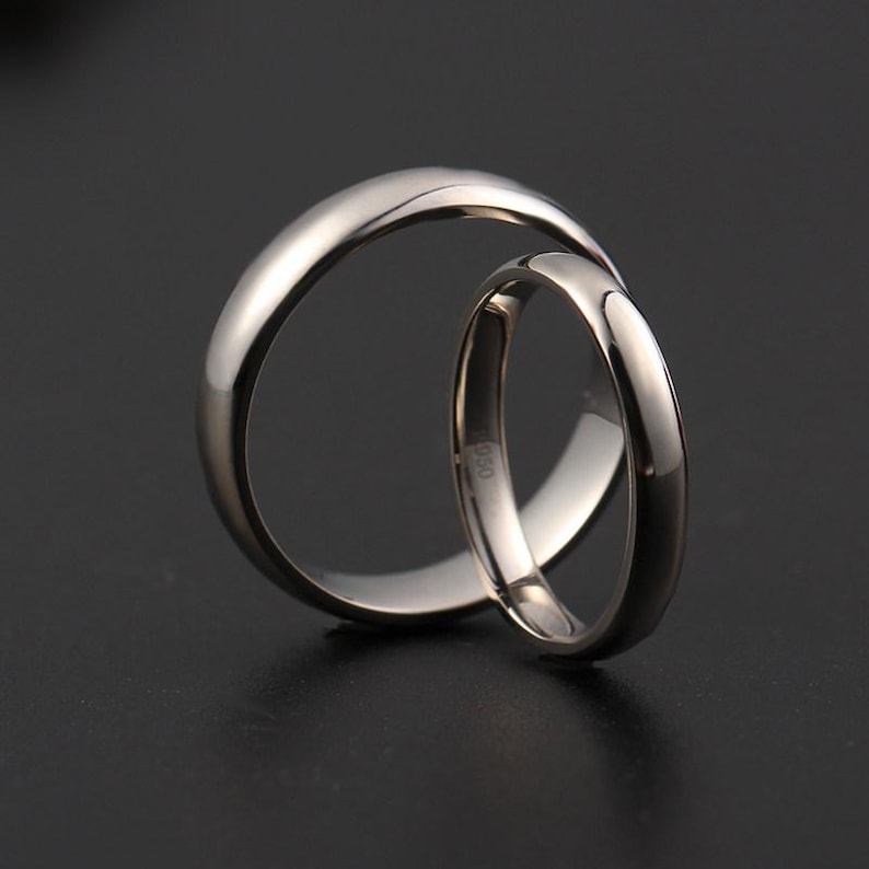 May include: Two silver wedding bands, one larger and one smaller, interlocked against a dark background. The rings have a polished, reflective surface. The smaller ring has the marking "950" on the inside.