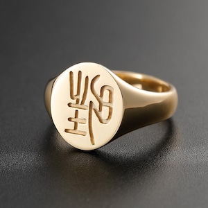 Puede incluir: Un anillo de sello dorado con un diseño grabado en una cara circular. El anillo tiene un acabado pulido y una banda gruesa. El diseño grabado presenta caracteres estilizados. El anillo está sobre un fondo oscuro.