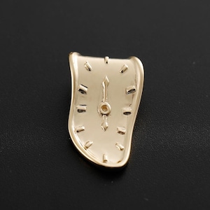 Puede incluir: Un broche de reloj dorado y derretido. El broche tiene un diseño clásico de esfera de reloj con marcadores y manecillas negras, pero la forma está distorsionada como si se derritiera. El broche está sobre una superficie negra.