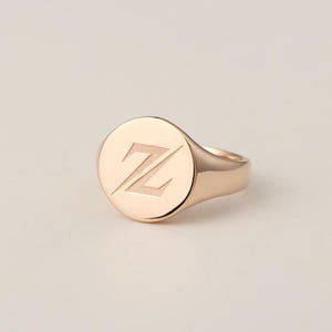 May include: A rose gold signet ring with a circular face. The face of the ring features the letter "Z" engraved in a stylized font. The ring is set against a neutral background.