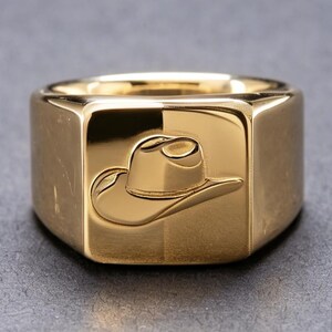 May include: A polished gold-tone signet ring featuring a square face with a debossed cowboy hat design. The ring has a smooth, reflective surface and a wide, solid band construction, showcasing a clean and metallic finish throughout the piece.