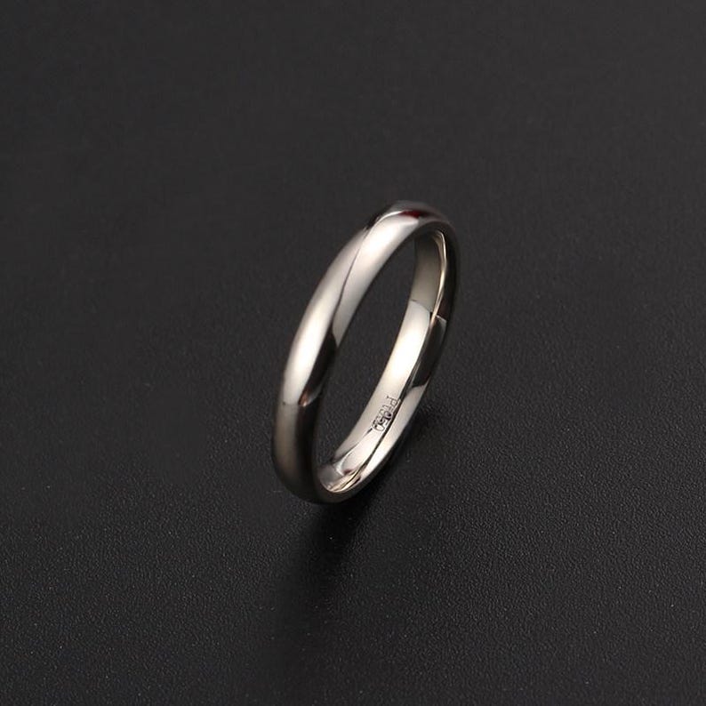 May include: A polished, silver-colored ring with a smooth, rounded band. The ring is set against a dark background, highlighting its reflective surface. The ring appears to be a wedding band or a simple, elegant piece of jewelry.