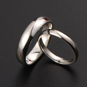 May include: Two polished silver rings, one wider than the other, are displayed against a dark background. The rings have a smooth, reflective surface, catching the light and creating highlights. The design is simple and elegant.