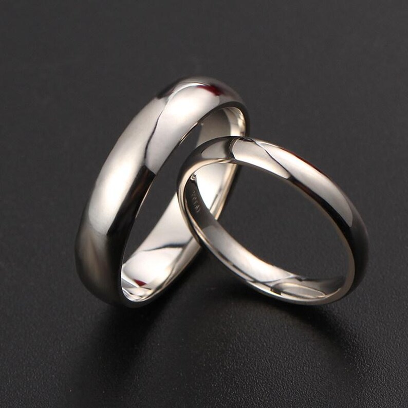 May include: Two polished silver rings, one wider than the other, are displayed against a dark background. The rings have a smooth, reflective surface, catching the light and creating highlights. The design is simple and elegant.