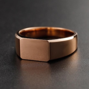 May include: A rose gold signet ring with a rectangular face. The ring has a polished finish and a simple, modern design. The ring is shown on a dark surface.