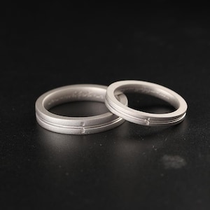 May include: Two silver rings with a matte finish are shown against a black backdrop. Each ring features a cross design and a horizontal line etched into the band. These rings are likely wedding bands or promise rings.