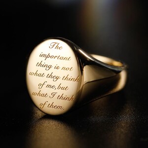 May include: A polished gold signet ring featuring a round face engraved with the text: The important thing is not what they think of me, but what I think of them. The ring has a smooth, reflective metallic surface and a classic band design.