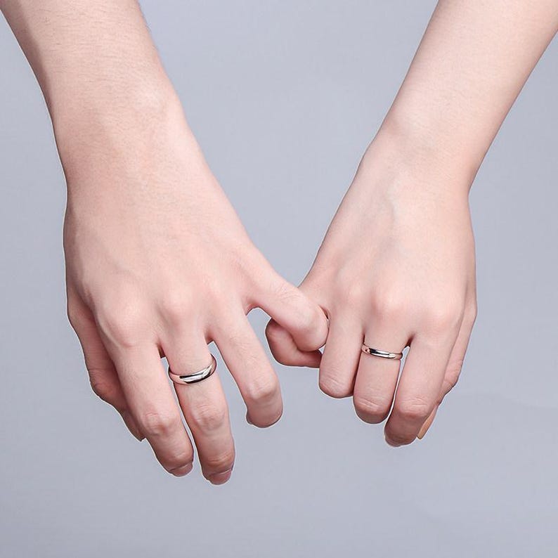 May include: Two hands holding each other, each wearing a silver ring. The rings are simple bands with a polished finish. The background is a soft, neutral color, highlighting the rings and the connection between the hands.