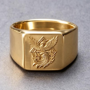 May include: A gold-toned signet ring featuring a flat, square face with an engraved design of a phoenix rising from flames within a geometric triangle frame. The band is wide and smooth with a polished finish, showcasing a detailed relief.