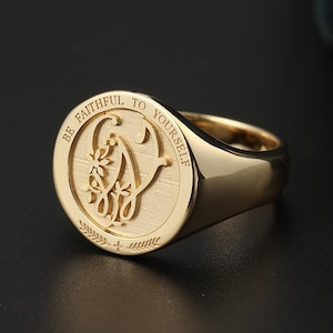 Puede incluir: Anillo de sello dorado con la inscripción "BE FAITHFUL TO YOURSELF" en el borde. El anillo presenta una letra "G" estilizada con detalles florales en el centro. El anillo está muy pulido y tiene una banda lisa y redondeada.