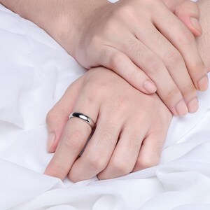 May include: A silver ring with a smooth, polished surface is worn on a finger. The ring has a simple, classic design. The background is a white, textured fabric.