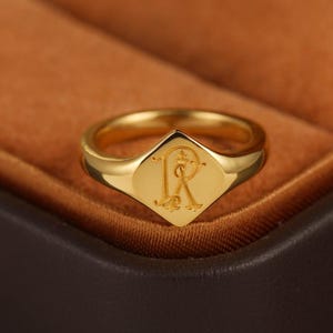 Könnte beinhalten: Ein goldfarbener Siegelring mit polierter Oberfläche und rautenförmiger Front. In die Mitte des Rings ist ein dekorativer, verzierter Buchstabe R in einer klassischen Serifenschrift eingraviert, der mittig auf der glatten, flachen Fläche liegt.