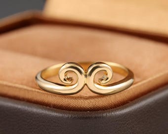May include: A gold-colored ring with a unique design. The ring features a central, stylized swirl motif. The ring is set against a brown background, suggesting a jewelry box or display.