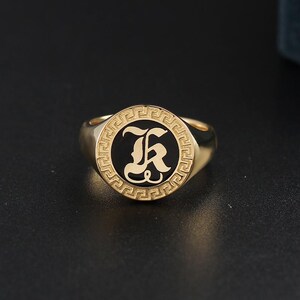 以下が含まれることがあります： 黒い中心に装飾的なフォントで「K」の文字が入った、金色のシグネットリング。 リングは、黒い中心の周りにギリシャキーのボーダーデザインがあります。 リングは光沢のある反射性の金属で作られています。