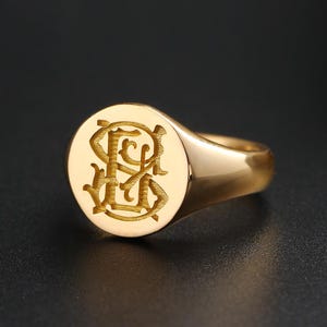 Può includere: Un anello con sigillo d'oro con finitura lucida. L'anello presenta un design con monogramma inciso su una faccia circolare. L'anello è posto su uno sfondo scuro, evidenziando la sua superficie riflettente.