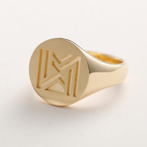 May include: A gold signet ring with a geometric design. The ring has a polished finish and a round face with an engraved pattern. The band is smooth and the ring is set against a white background.