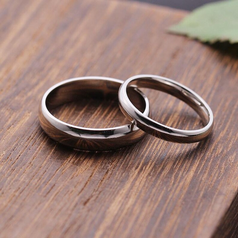 May include: Two silver wedding rings, one larger and one smaller, are displayed on a wooden surface. The rings have a polished, reflective finish, and the larger ring has a wider band. The background is a warm, natural wood grain.