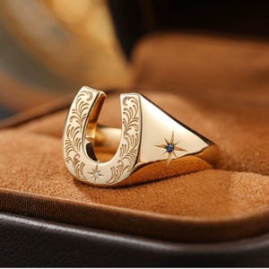 Könnte beinhalten: Ein goldfarbener Ring mit Hufeisen-Design, mit aufwendigen floralen Gravuren und einem Stern mit blauem Edelstein. Der Ring ist vor einem braunen Hintergrund platziert.
