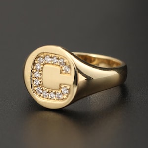 Puede incluir: Un anillo de sello dorado con la letra "C" incrustada con pequeñas piedras preciosas transparentes. El anillo tiene una superficie pulida y reflectante y una cara redondeada. La banda es lisa y se estrecha hacia la parte posterior.