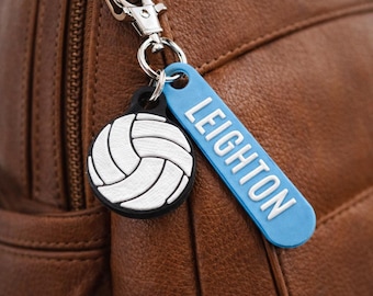 May include: A volleyball keychain with a white and black design, and a blue tag that says "LEIGHTON" in white letters. The keychain is attached to a silver clasp, and is set against a brown leather background.