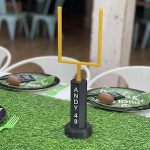 Può includere: Un mini-palo da football con montanti gialli e base nera. La base riporta il nome "ANDY" e il numero "49". Il palo è posto su una superficie verde.