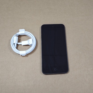 May include: A black smartphone with a rectangular screen and a circular home button at the bottom, accompanied by a white charging cable coiled into a circle with a standard connector plug at one end and a device connector at the other end.
