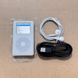 May include: A white digital music player with a circular navigation wheel and a screen displaying a menu with options for Music, Photos, Extras, Settings, Shuffle Songs, Backlight, and Now Playing, accompanied by white wired earbuds and a black USB cable.