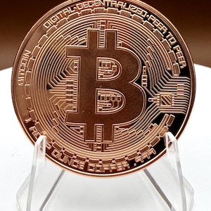 Puede incluir: Una moneda Bitcoin de cobre exhibida en un soporte transparente. La moneda presenta el símbolo de Bitcoin, intrincados patrones similares a circuitos y las palabras "DIGITAL DECENTRALIZED PEER TO PEER" y "1 OUNCE COPPER .999".