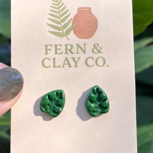 May include: A pair of leaf-shaped, dark green earrings on a cream-colored card. The card features a fern and a vase illustration, with the text "FERN & CLAY CO." printed in green. The earrings are approximately 0.75 inches in length.